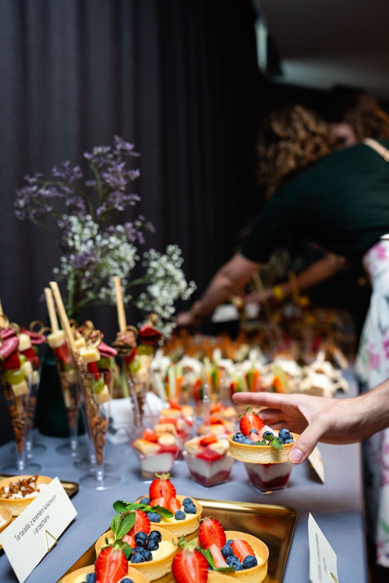 Stół cateringowy z tartaletkami z kremem, owocami (truskawki, borówki) i przekąskami w szklankach. W tle osoba przygotowuje ofertę 'Luxury'.