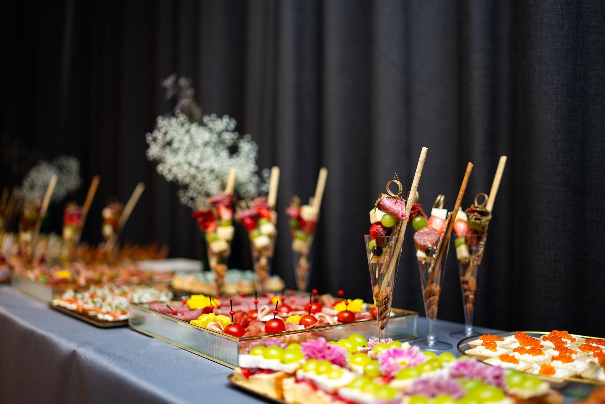 Luksusowy catering: elegancko podane przekąski na stole z szarego obrusa, w szklanych naczyniach i na paterach, z dekoracją kwiatową na ciemnym tle.