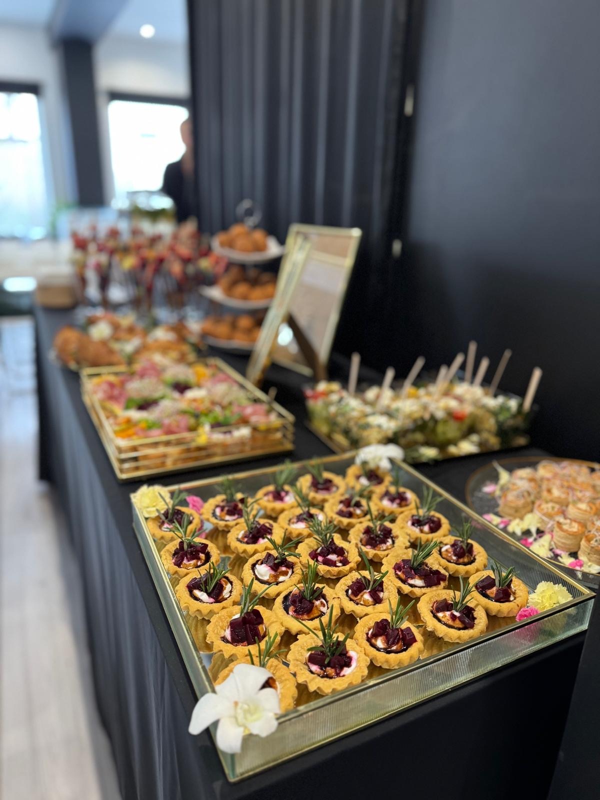 Elegancki stół cateringowy z tartaletkami z burakiem i rozmarynem na pierwszym planie. W tle widoczne inne przekąski i dekoracje, tworzące luksusową aranżację.