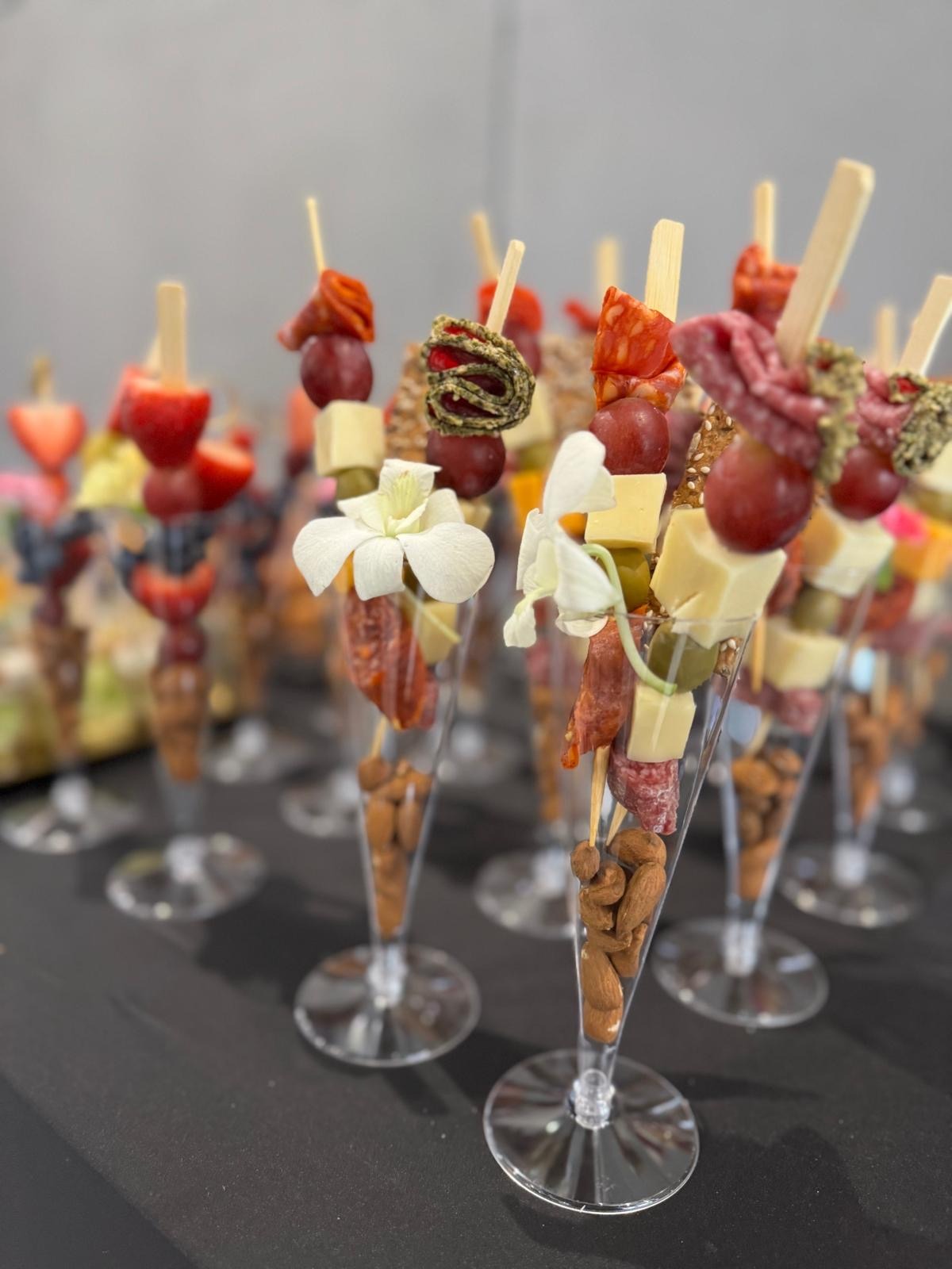 Ekskluzywne finger food w kieliszkach: migdały, ser, winogrona, salami, dekoracja kwiatowa. Elegancki catering na ciemnym stole, w tle inne przekąski.