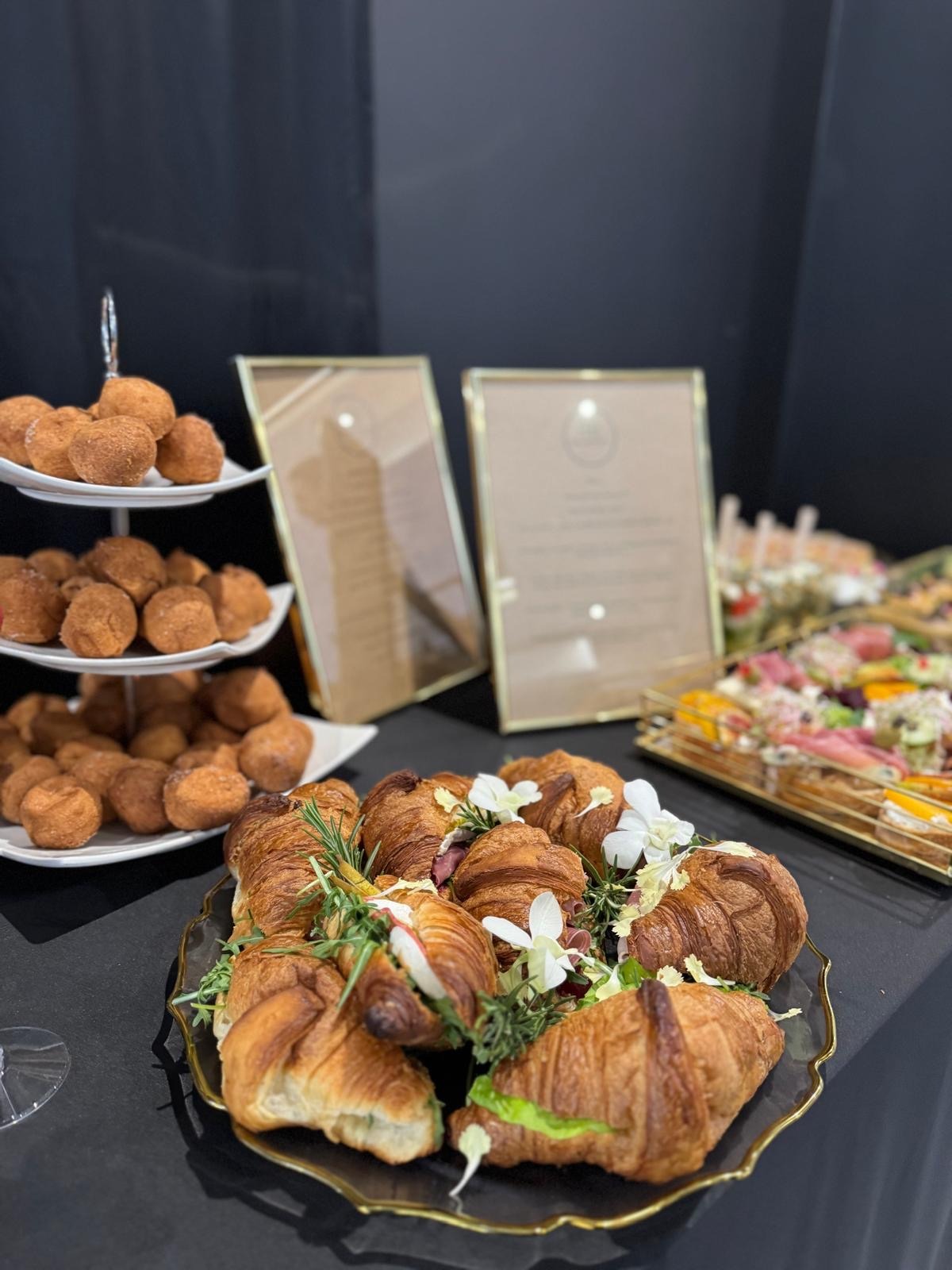 Elegancki catering: kruche croissanty z nadzieniem i kwiatami na złotej tacy, obok pączki na etażerce i kanapeczki. W tle menu w ramkach.