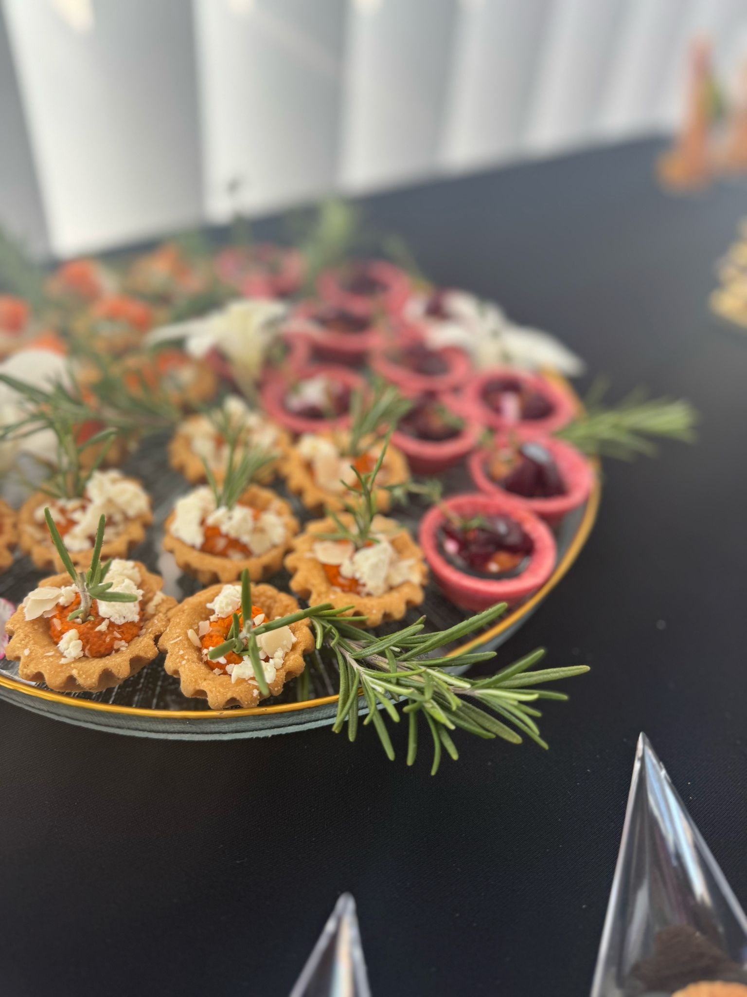 Elegancki catering: tartaletki z farszem, serem i rozmarynem na talerzu z czarnym tłem, część innych przekąsek widoczna w tle. Luksusowa oferta.