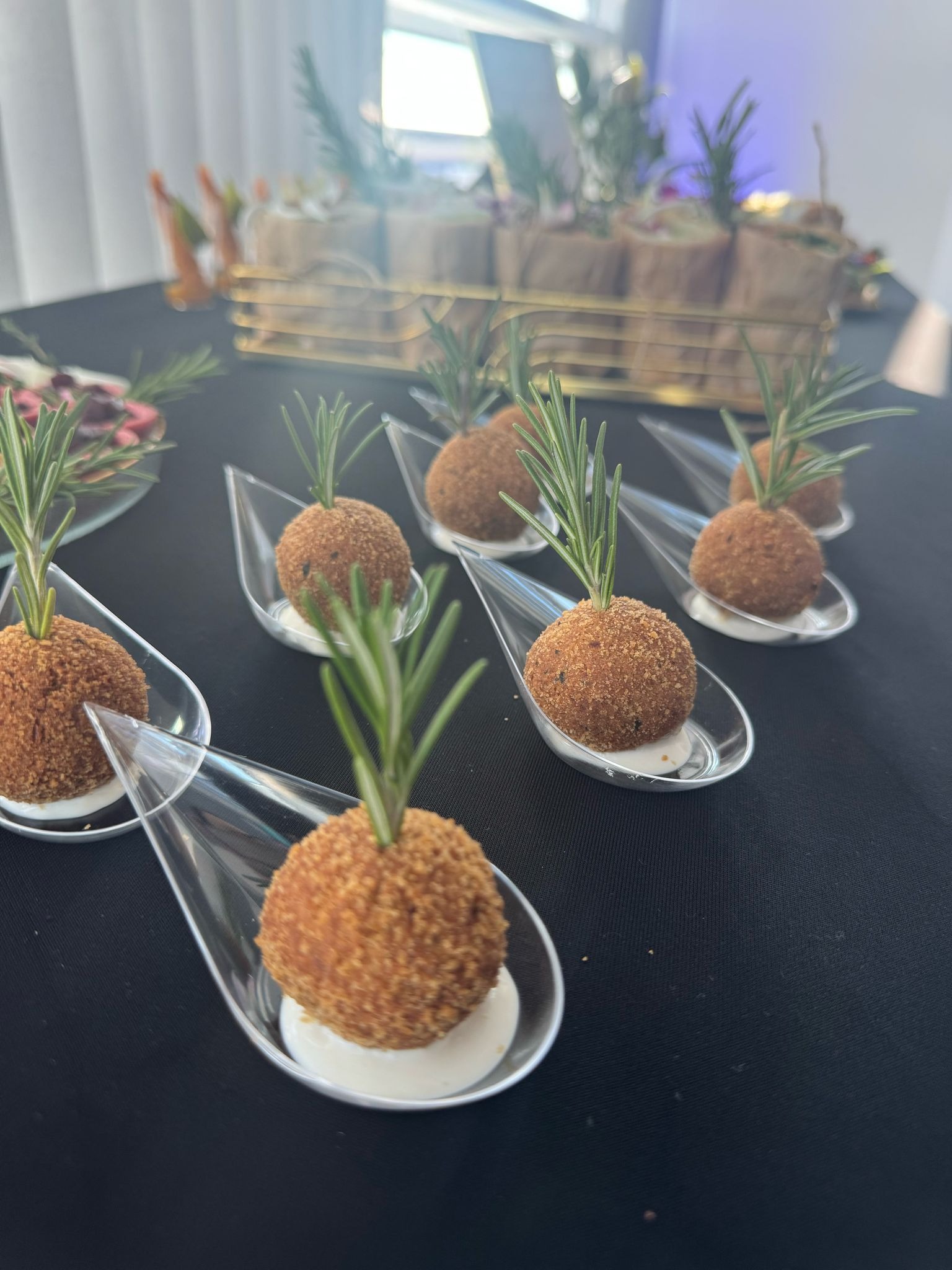 Eleganckie przekąski cateringowe: kulki w panierce z rozmarynem, podane na przezroczystych łyżeczkach na czarnym stole. W tle inne dania.