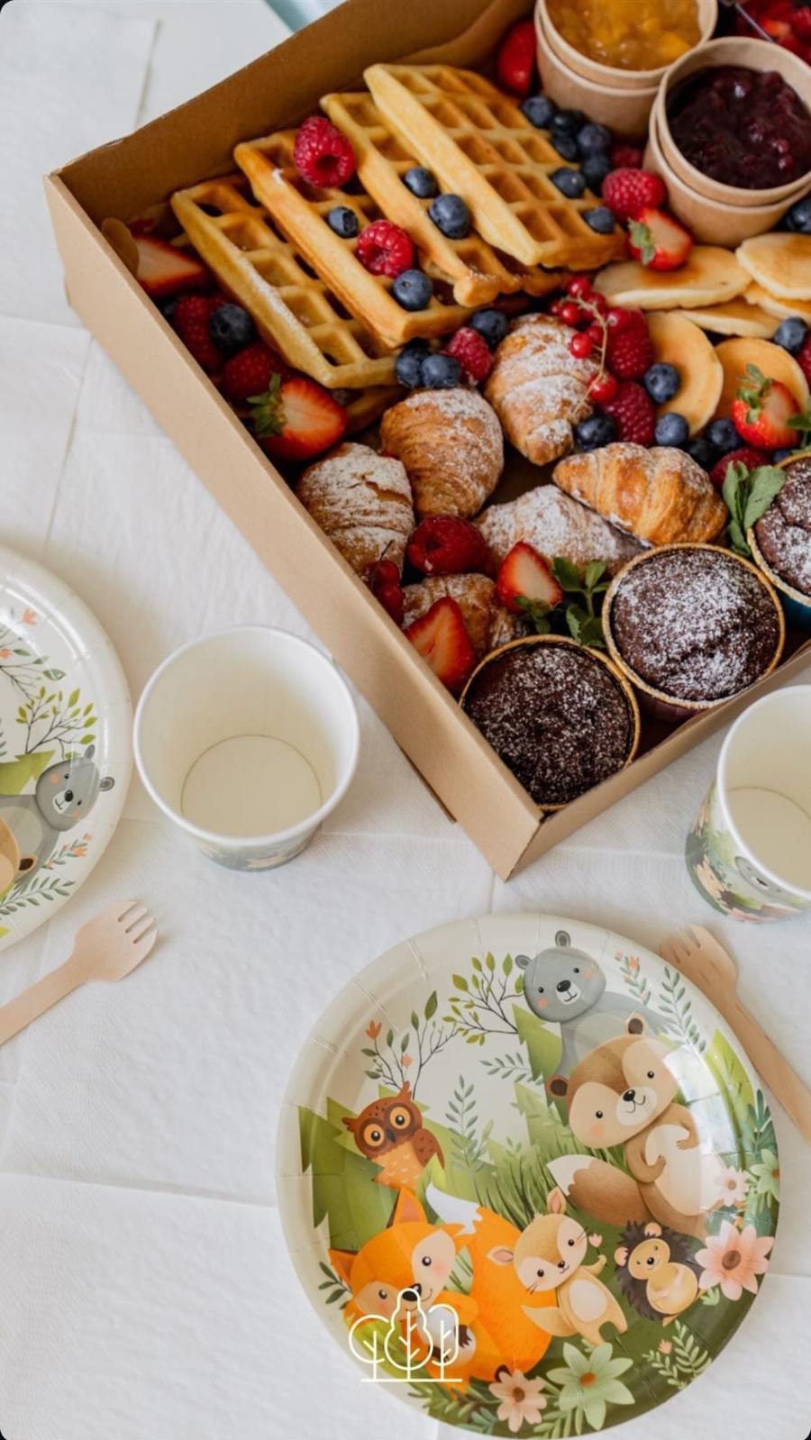 Box z cateringiem: gofry, croissanty, muffiny i owoce, obok talerzyki i kubeczki z motywem leśnych zwierząt, tworzące zestaw na przyjęcie dla dzieci.