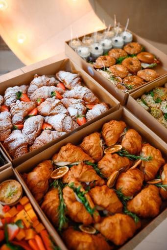 Kartonowe boxy z różnorodnym cateringiem: słodkie rogaliki z pudrem i truskawkami, mini-deserki w kubeczkach, kanapki z sezamem i rogaliki z dodatkami.