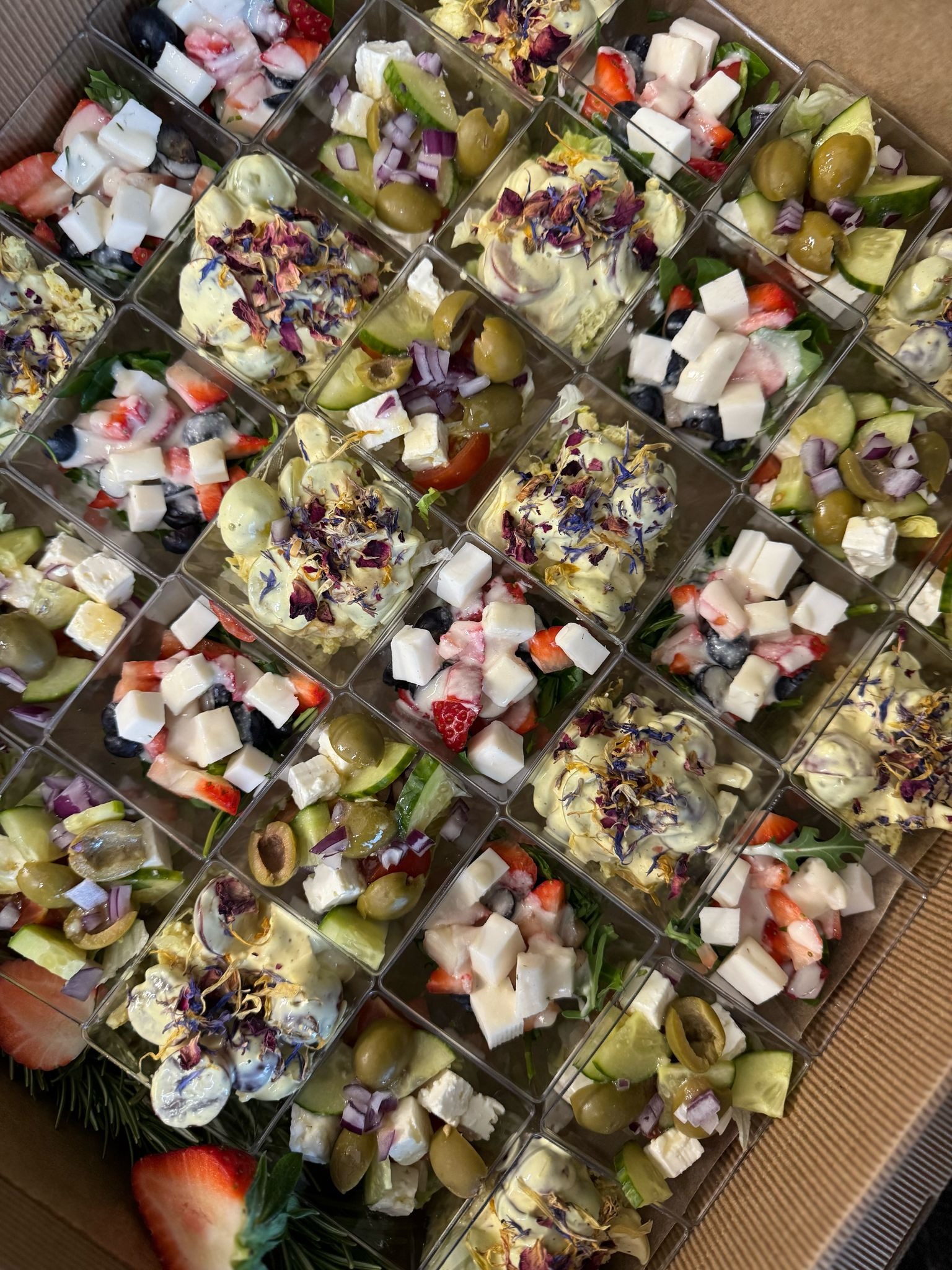 Catering w boxach: różnorodne sałatki w przezroczystych pojemnikach, widoczne świeże składniki, np. truskawki, feta, oliwki, dekorowane jadalnymi kwiatami.