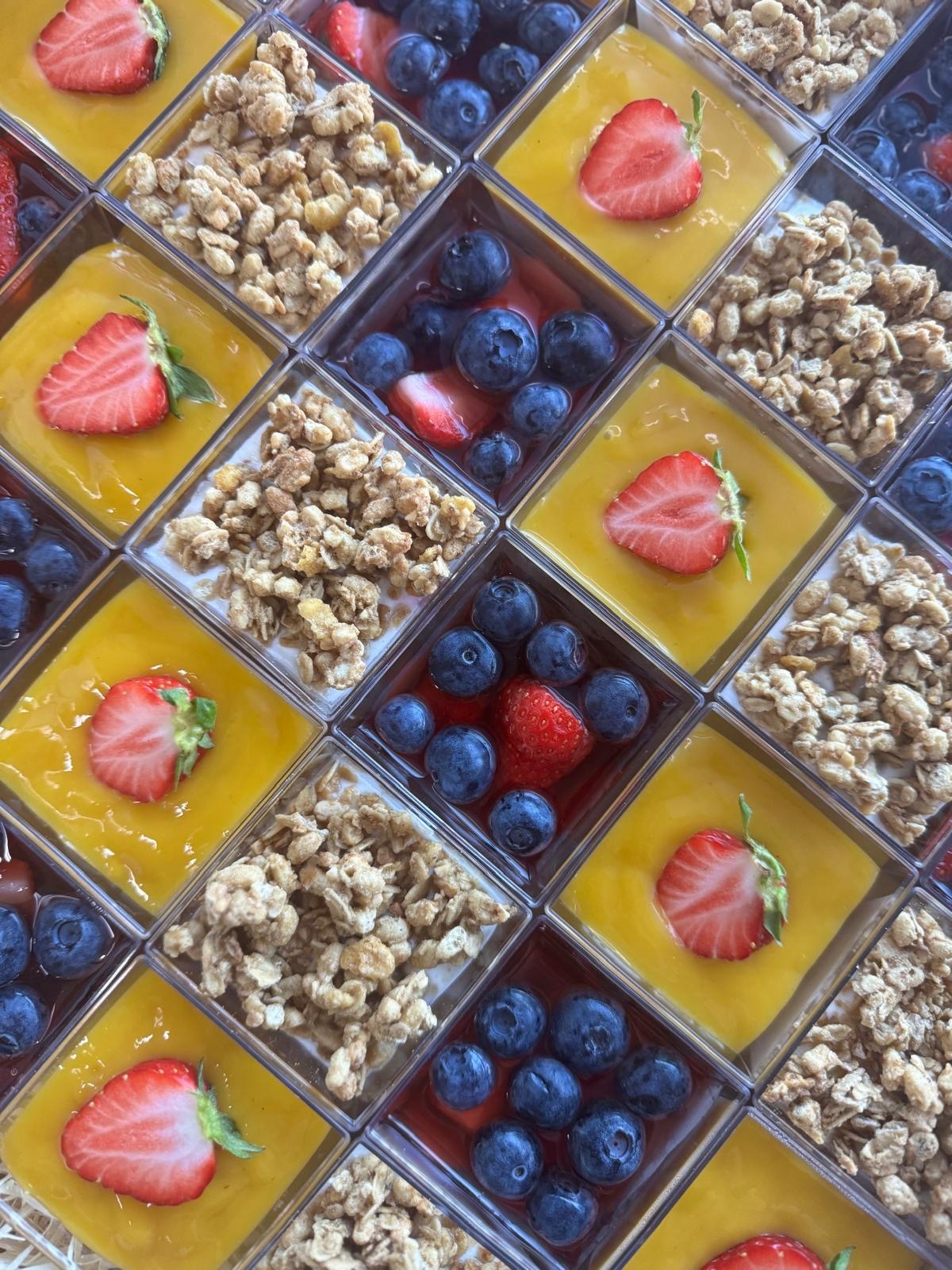 Boxy cateringowe: desery z owocami (truskawki, borówki) i granolą, ułożone w kwadratową mozaikę. Żółty krem, widok z góry, świeże składniki.