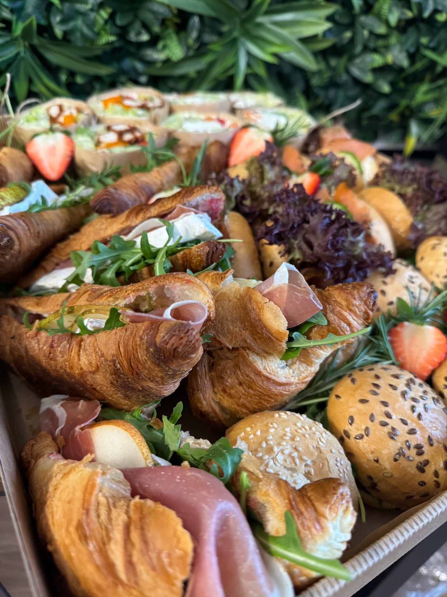 Apetyczny catering w boxie: croissanty z szynką, roladki z warzywami i bułeczki, wszystko udekorowane świeżymi truskawkami i zielenią na tle dekoracyjnej ściany.