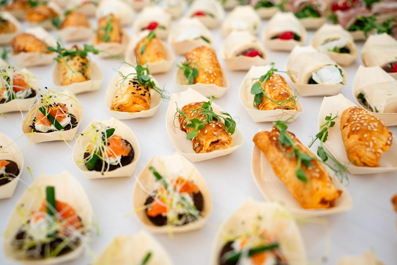Różnorodne, eleganckie przekąski w małych, drewnianych łódeczkach, ułożone rzędami na białym obrusie. Widoczne roladki, tartinki z łososiem i mini tortille.
