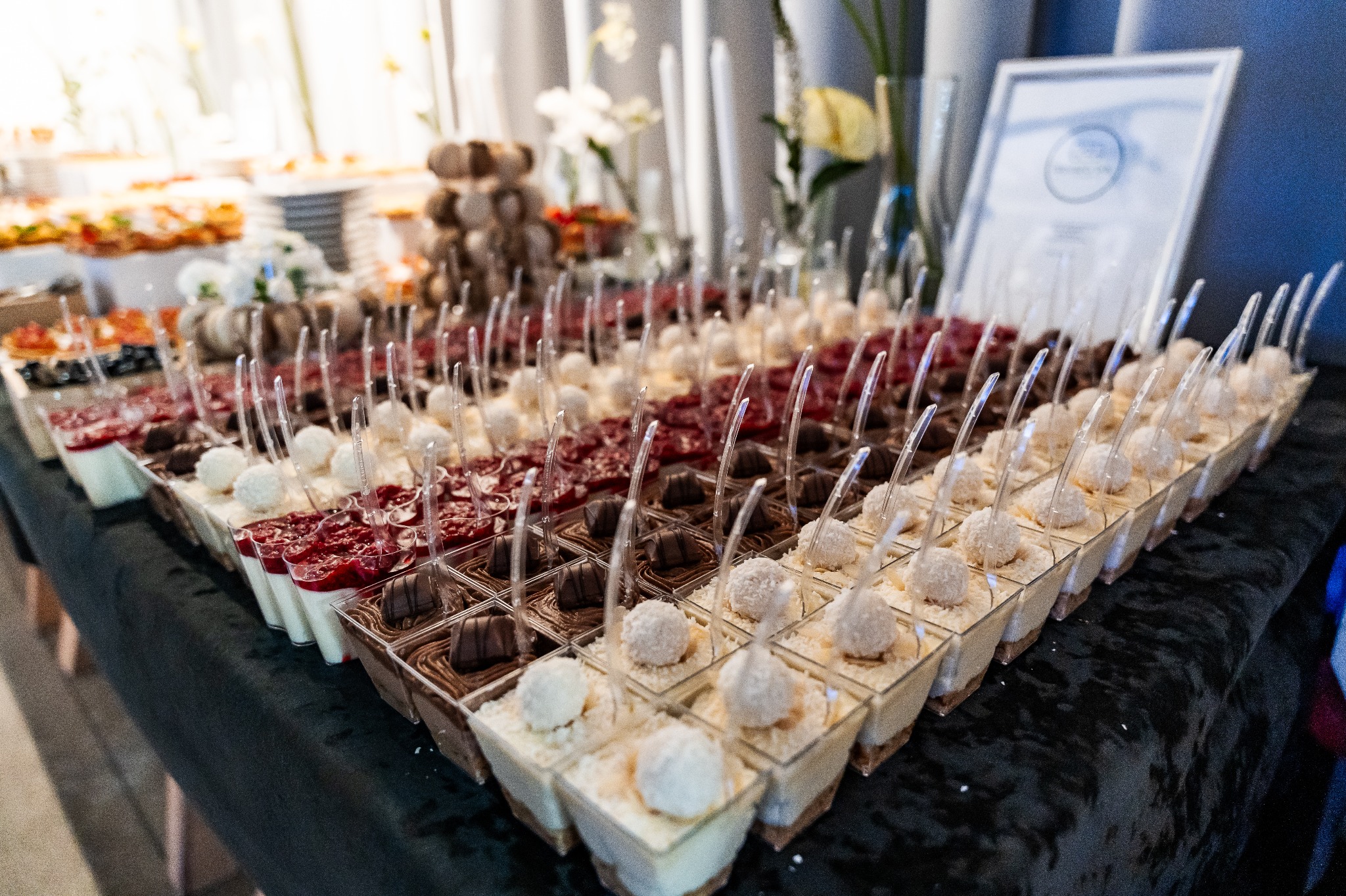 Rzędy deserów w małych pojemnikach z łyżeczkami, różnorodne smaki: kokosowe, czekoladowe, z czerwoną galaretką. Bufet słodki na przyjęciu, widok z góry.