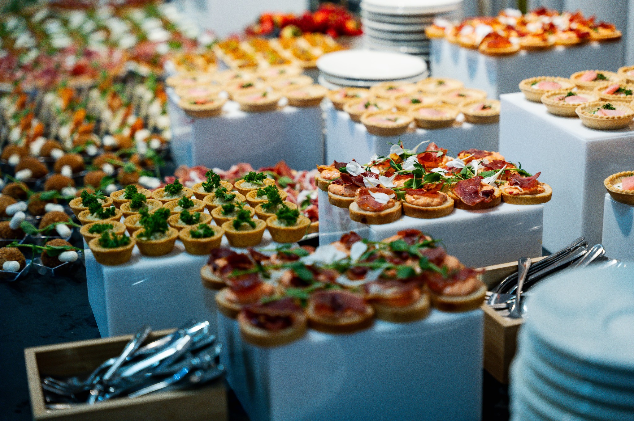 Elegancki catering: tartaletki z ziołami, mini-kanapeczki z szynką i kwiatami, kulki mięsne na białych podestach. W tle kwiaty i stosy talerzy.