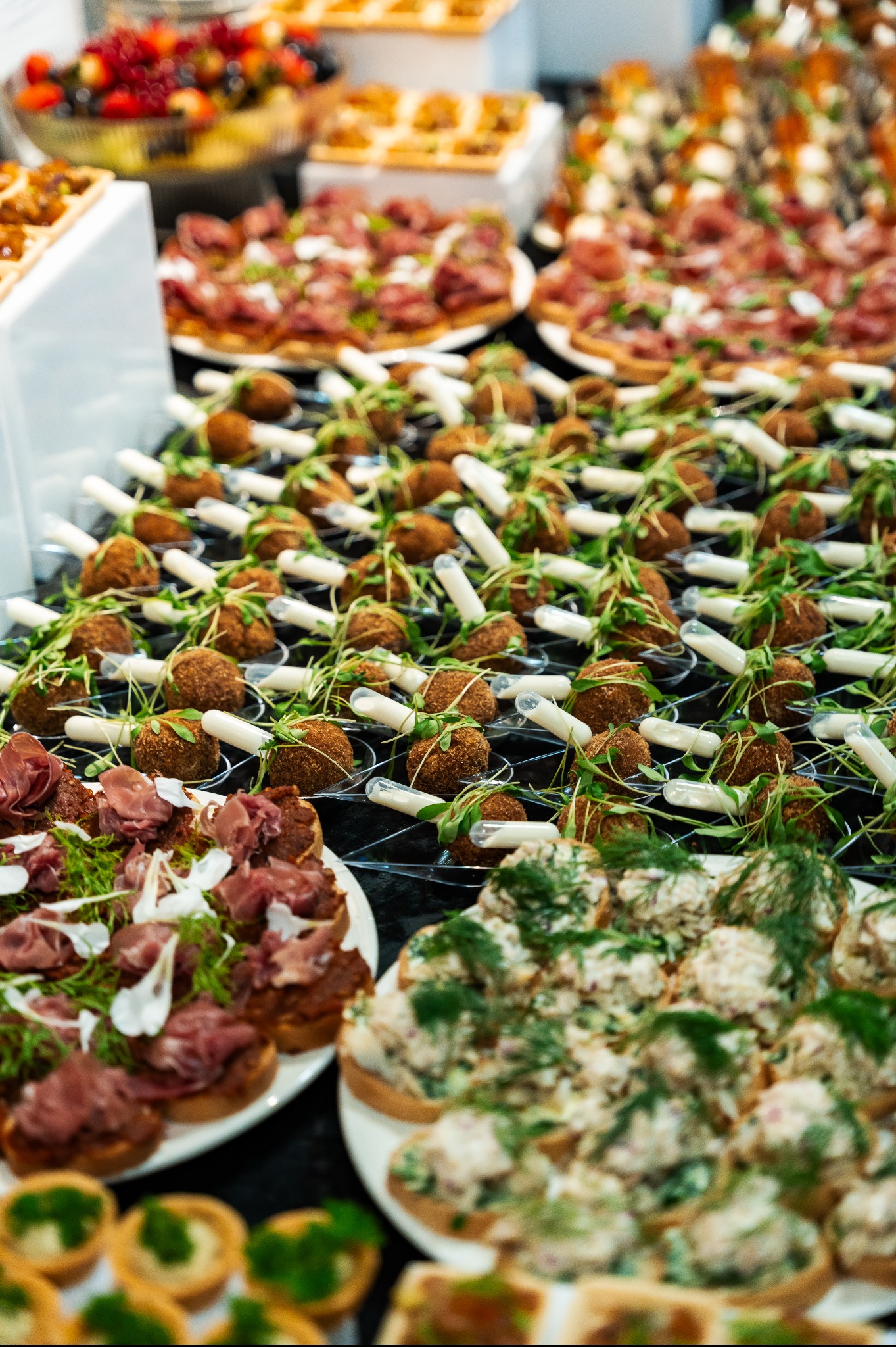 Bogaty wybór przekąsek cateringowych: mini tartaletki, kanapeczki z pastami, koreczki z mięsem i kulki z zielonymi dodatkami, ułożone na czarnym stole.