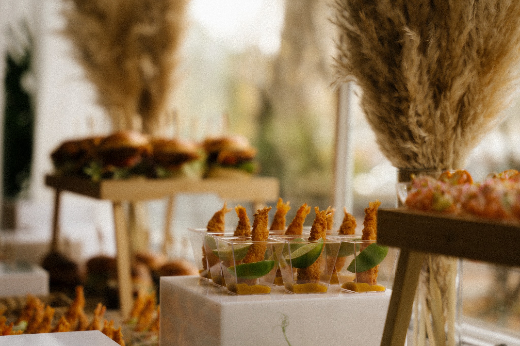 Rustykalny catering w Poznaniu: mini burgery i krewetki w tempurze z limonką, elegancko podane na drewnianych stojakach. Dekoracja z traw pampasowych w tle.