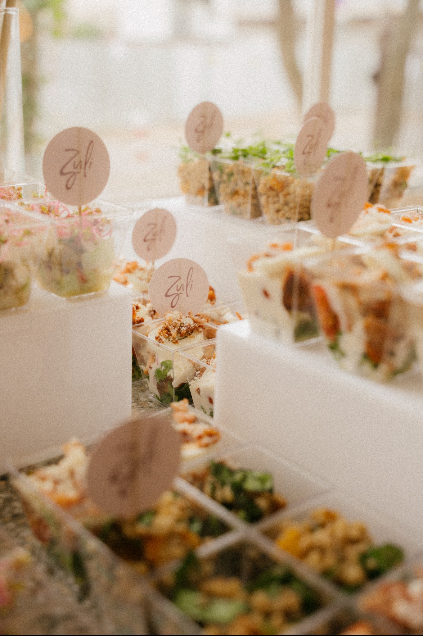 Elegancki catering w przezroczystych pojemnikach z logo 'Zuli' na beżowych etykietach. Różnorodne sałatki i przekąski na białym tle, delikatne oświetlenie.