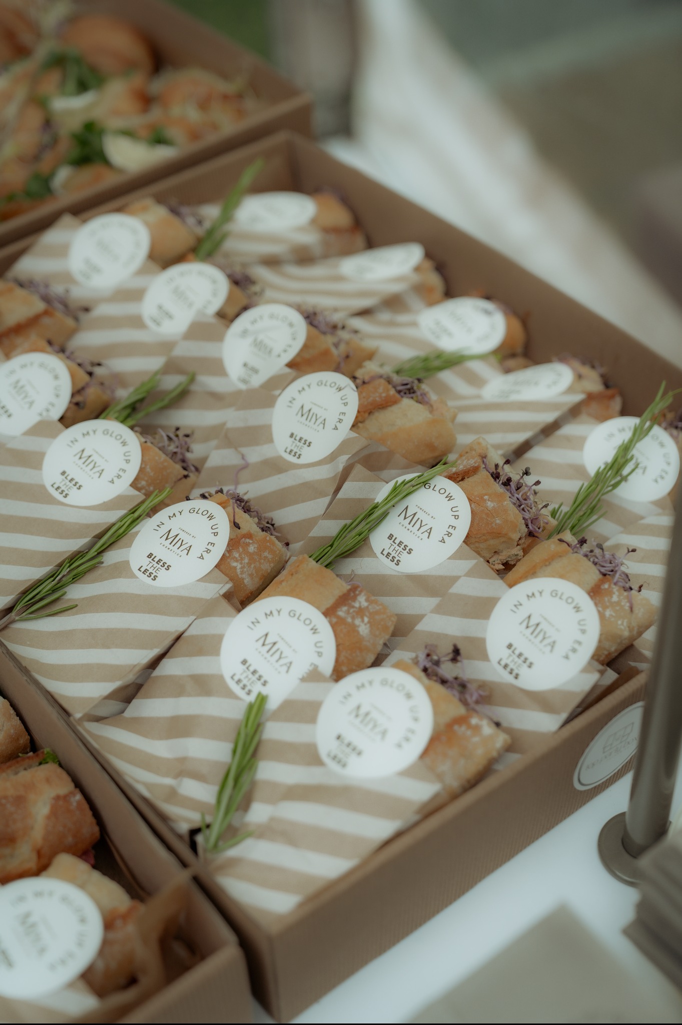 Personalizowane kanapki w pudełkach z logo Miya Cosmetics, udekorowane gałązkami rozmarynu i fioletowymi kwiatami. Elegancki catering firmowy na jasnym tle.