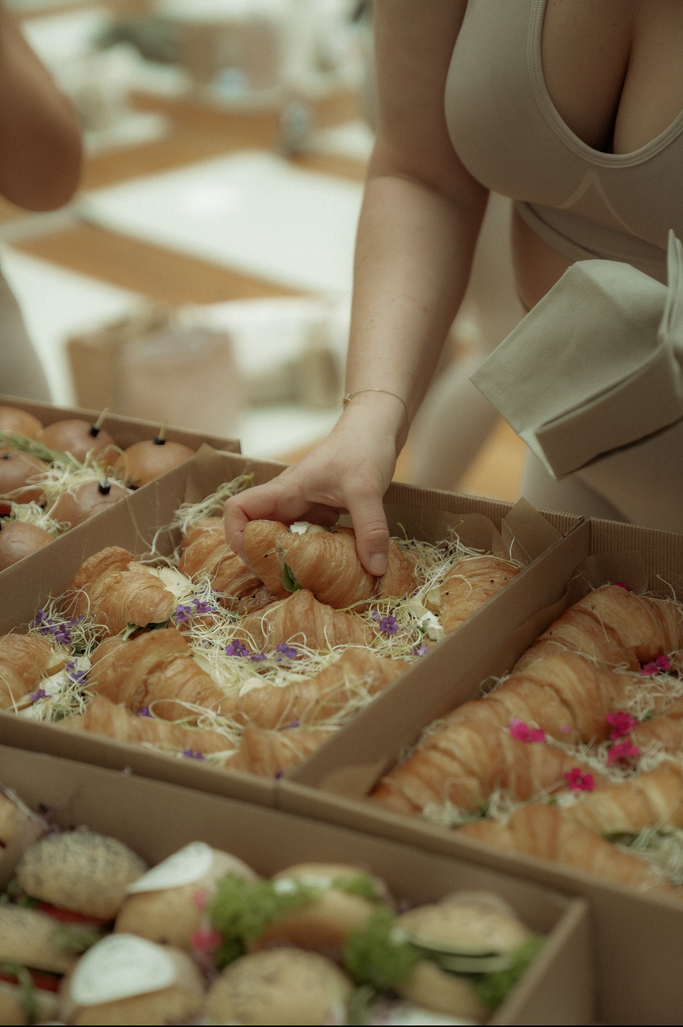 Kobieta wkłada rogalika do kartonowego pudełka z cateringiem: kanapki, rogaliki z dekoracją z kwiatów i ziół. Pudełka z jasnego kartonu.