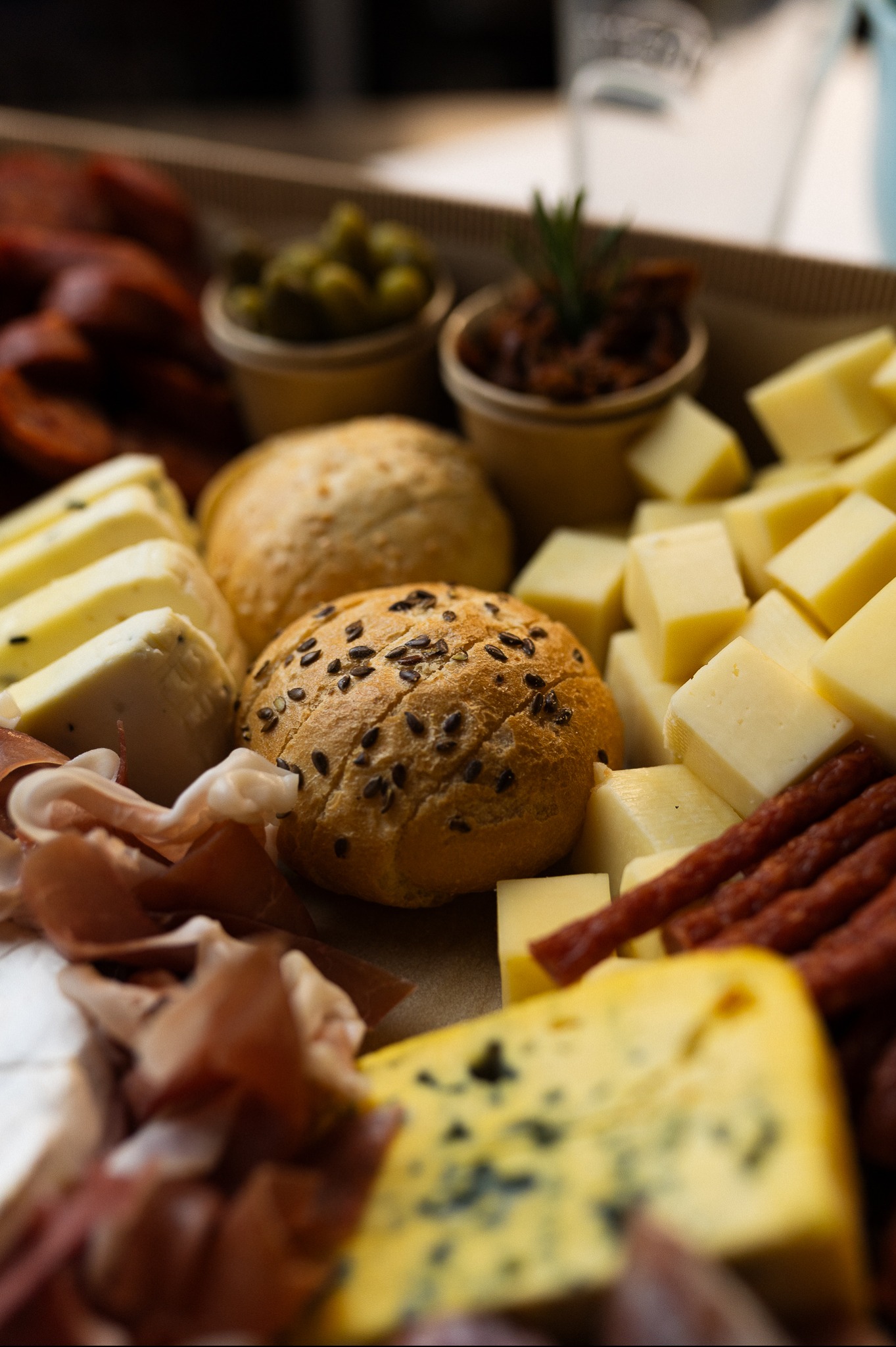 Box cateringowy: bułki z ziarnami, sery żółte i pleśniowe, wędliny, oliwki i suszone pomidory w kubeczkach. Całość w kartonowym pudełku.