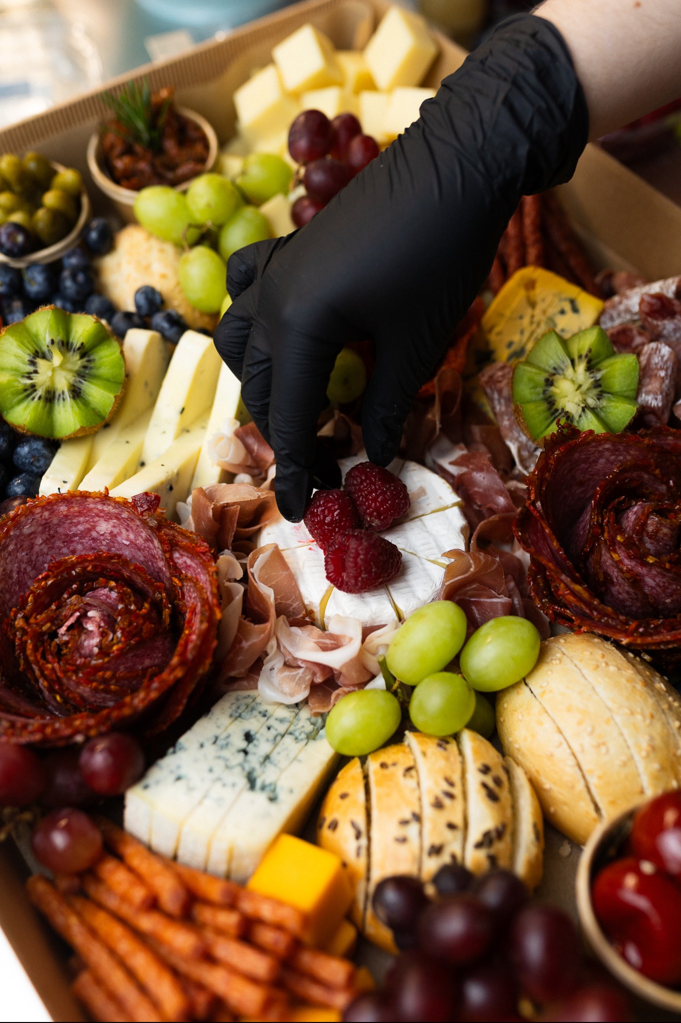 Elegancki box cateringowy: ręka w czarnej rękawiczce układa maliny na serze brie. W tle sery, wędliny, owoce, pieczywo i pikle, tworzące apetyczną kompozycję.