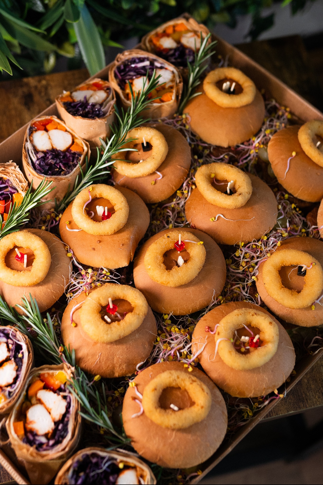 Box cateringowy: tortille z warzywami i kurczakiem oraz mini burgery z krążkami cebulowymi, dekorowane rozmarynem i kiełkami, na drewnianym tle.