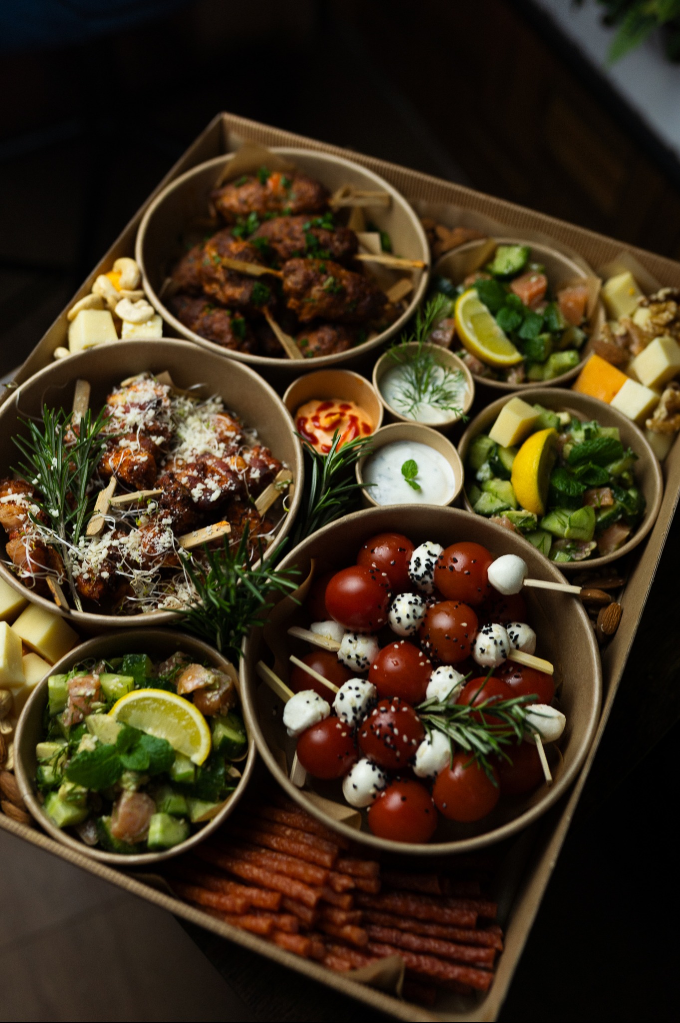 Box cateringowy z różnorodnymi przekąskami: szaszłyki z pomidorkami i mozzarellą, mięsne koreczki, sałatki warzywne, sery i sosy. Elegancka prezentacja w ekologicznych naczyniach.