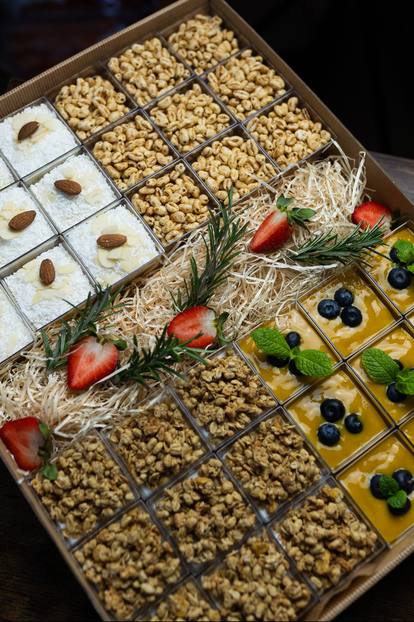 Catering w boxach: pudełko z przegródkami, w których znajdują się desery (kokosowe, z mango i borówkami, granola, prażony ryż) udekorowane owocami i rozmarynem.