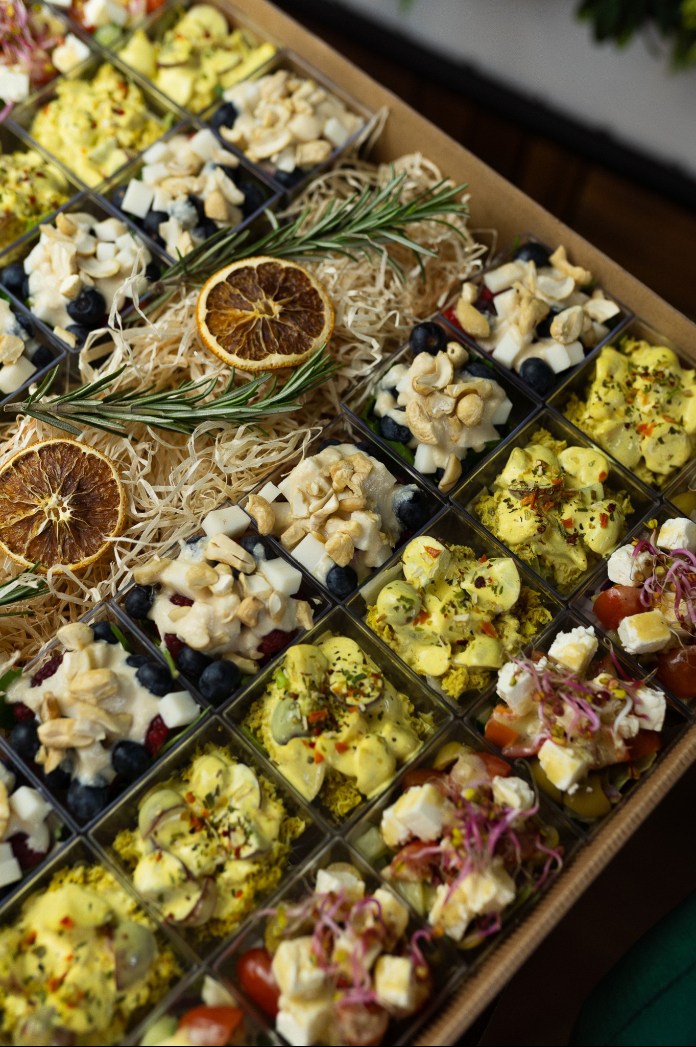 Box cateringowy z różnorodnymi przekąskami w przezroczystych pojemnikach, udekorowany suszonymi pomarańczami i gałązkami rozmarynu, wypełniony siankiem.