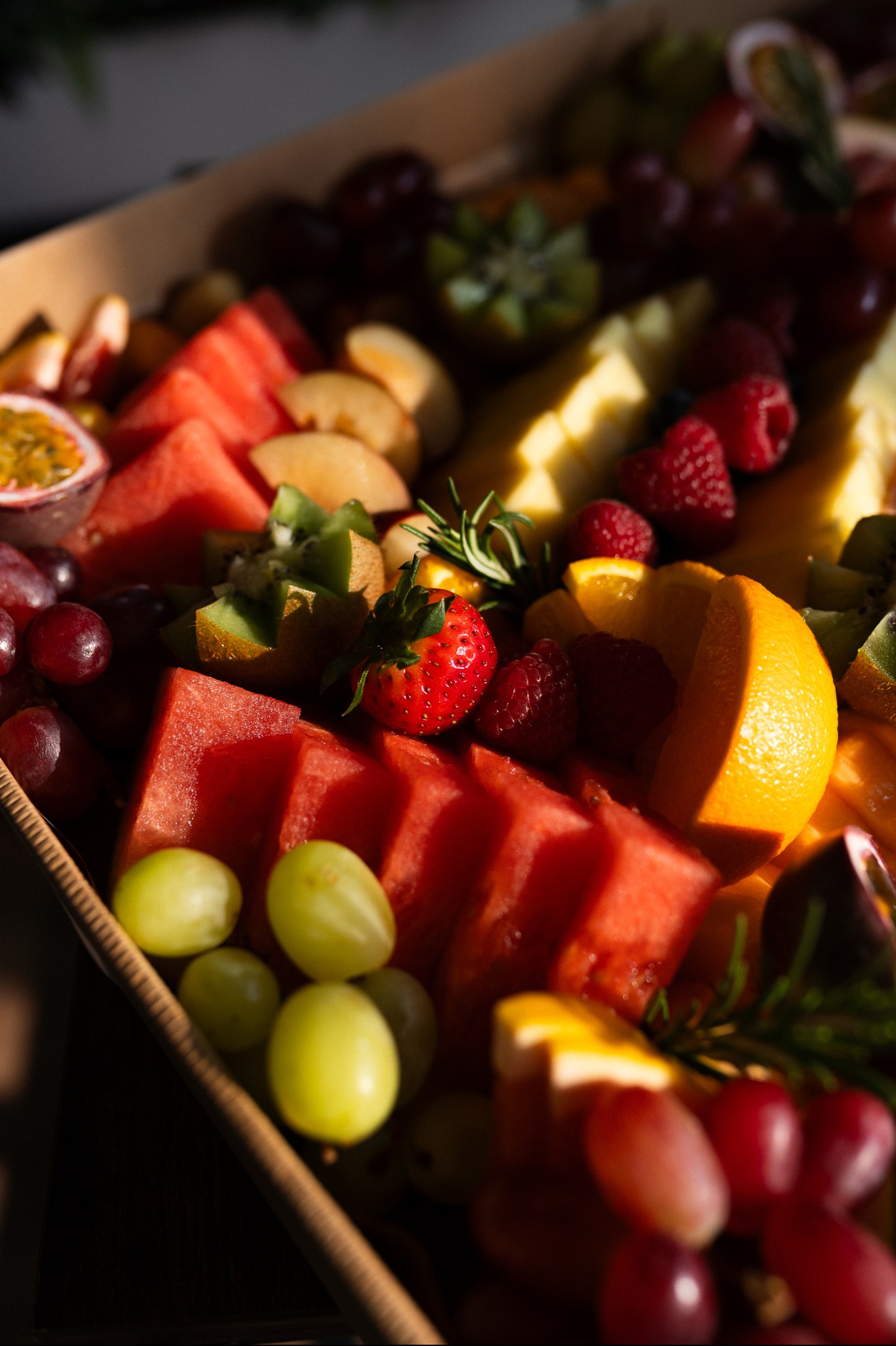 Box pełen świeżych owoców: arbuz, winogrona, kiwi, maliny i pomarańcze, skąpany w naturalnym świetle. Idealny na catering firmowy lub świąteczny.