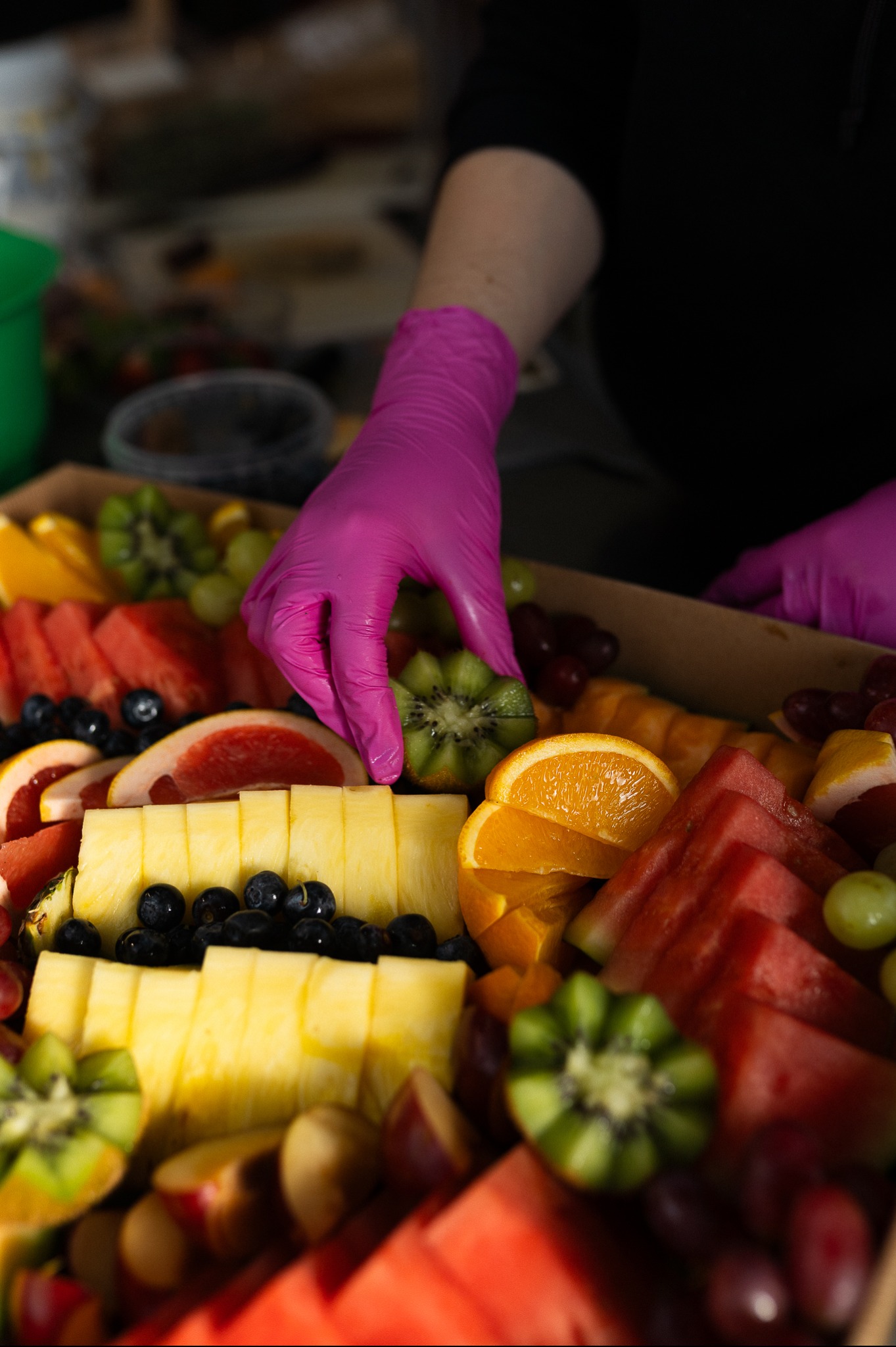 Ręka w różowej rękawiczce układa plaster kiwi w boxie z owocami: arbuz, ananas, pomarańcze, grejpfruty, winogrona i borówki. Klasyczny catering w boxach.