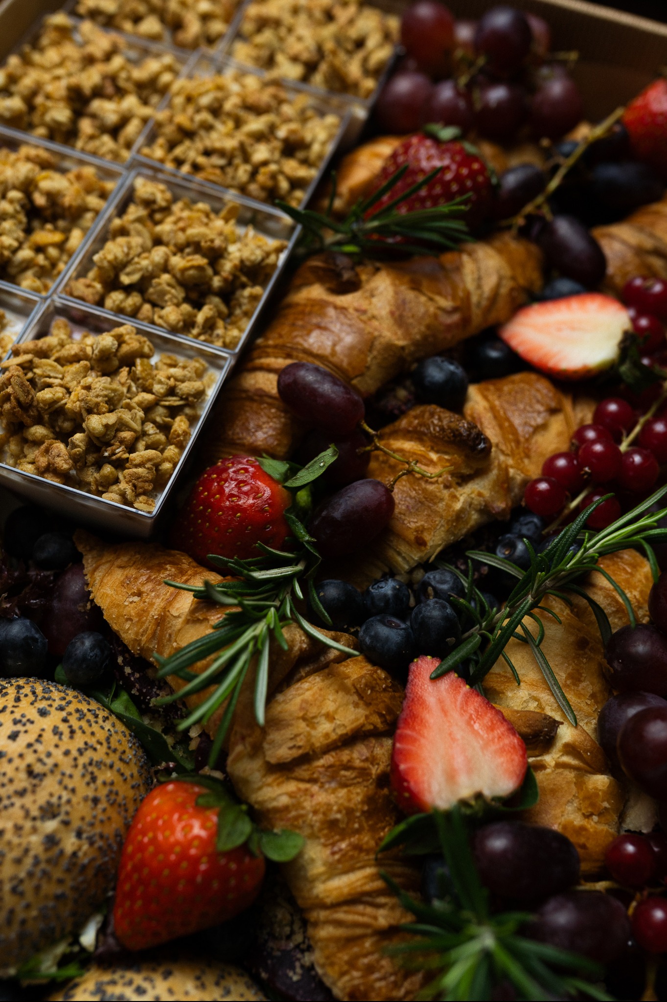 Bogata kompozycja cateringowa: rogaliki, owoce (winogrona, truskawki, jagody), granola w pojemnikach i gałązki rozmarynu tworzą apetyczną całość.