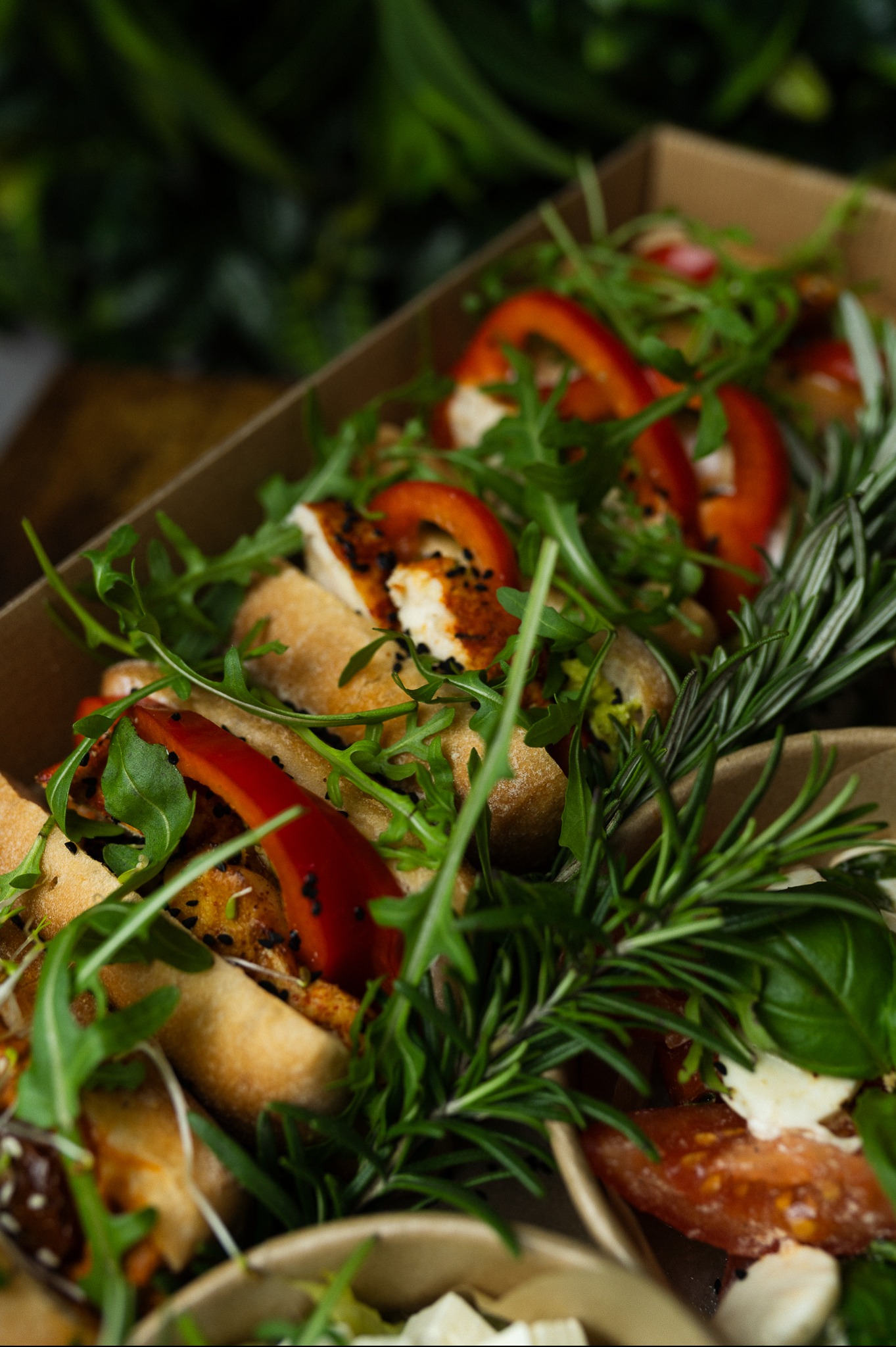 Kreatywnie zapakowany catering w boxach: kanapki z papryką, rukolą i rozmarynem, widoczne w brązowym pudełku na tle zieleni. Estetyczna kompozycja.