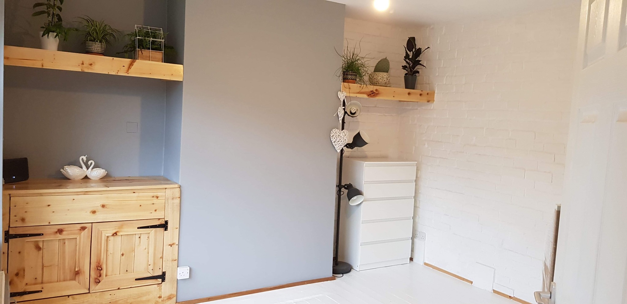 Wnętrze pokoju po remoncie: szara ściana kontrastuje z białą cegłą, drewniane półki z roślinami, komoda z surowego drewna i biała komoda z szufladami.