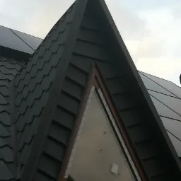 Szczegółowe ujęcie szczytu dachu z oknem trójkątnym w trakcie montażu, widoczne zabezpieczenia okna, ciemna blacha dachowa o falistej strukturze.