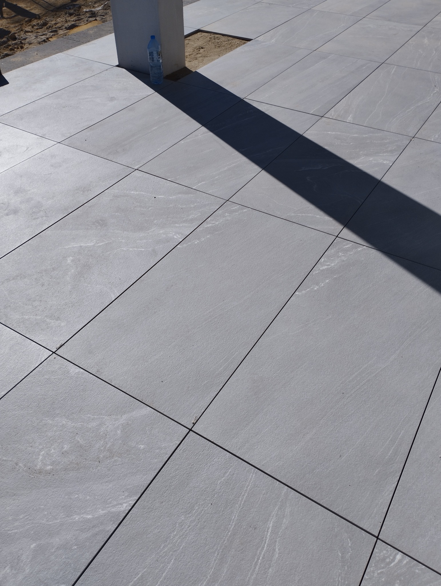 Ułożona szara, matowa posadzka z płytek ceramicznych na zewnątrz, widoczny cień słupa i butelka wody mineralnej. Płytki o delikatnej strukturze przypominającej kamień.