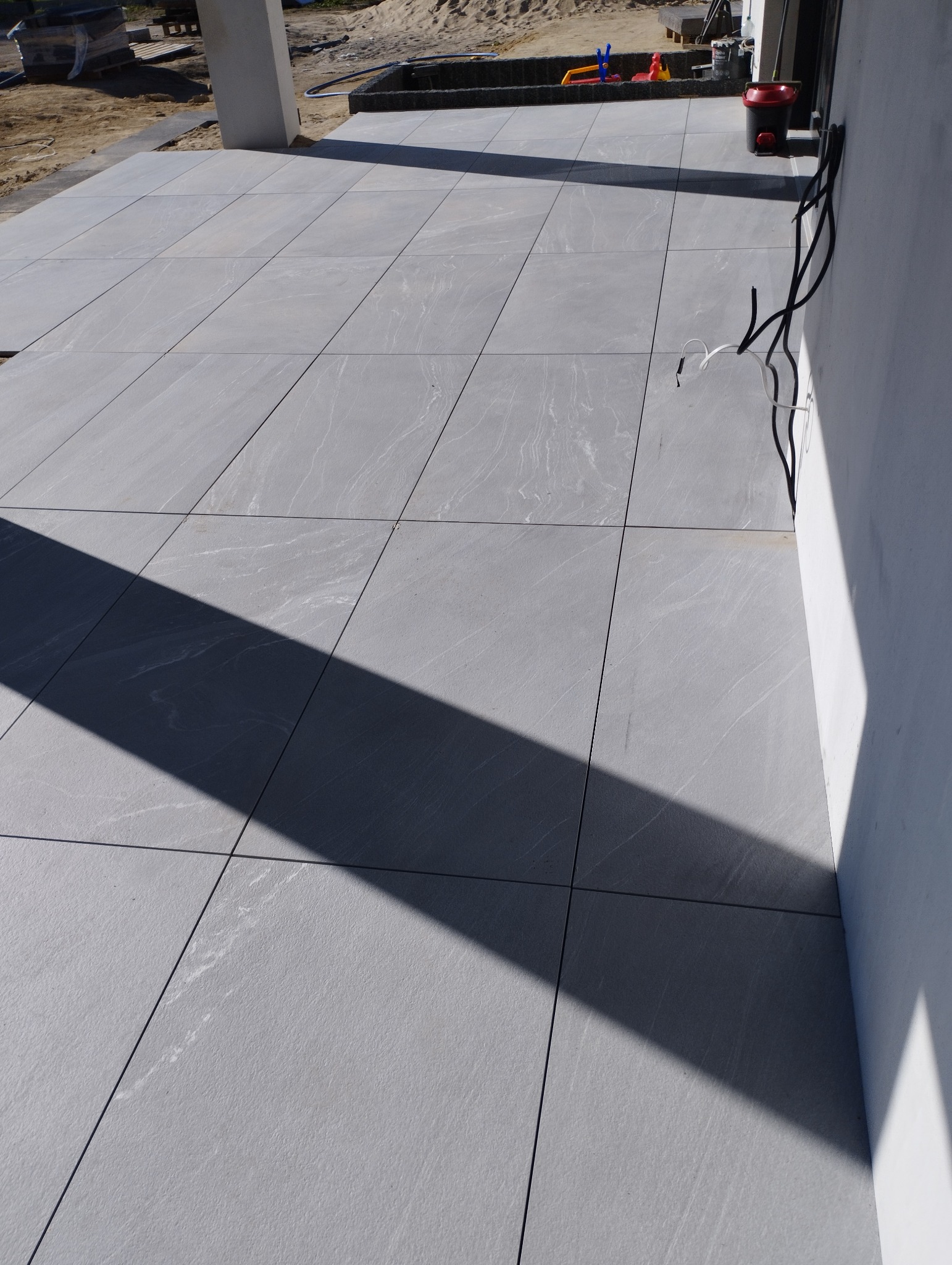 Szare płytki ułożone na zewnątrz, cień rzucany przez element budynku, widoczne przewody elektryczne przy ścianie budynku.