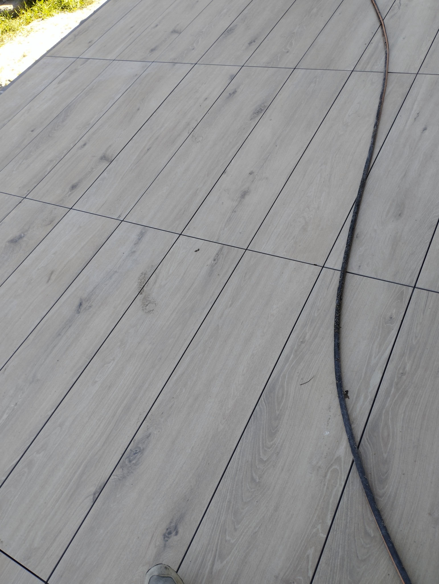 Ułożona pod kątem jasna podłoga z płytek imitujących drewno, widoczna czarna fuga i fragment brudnego kabla wzdłuż krawędzi płytek.