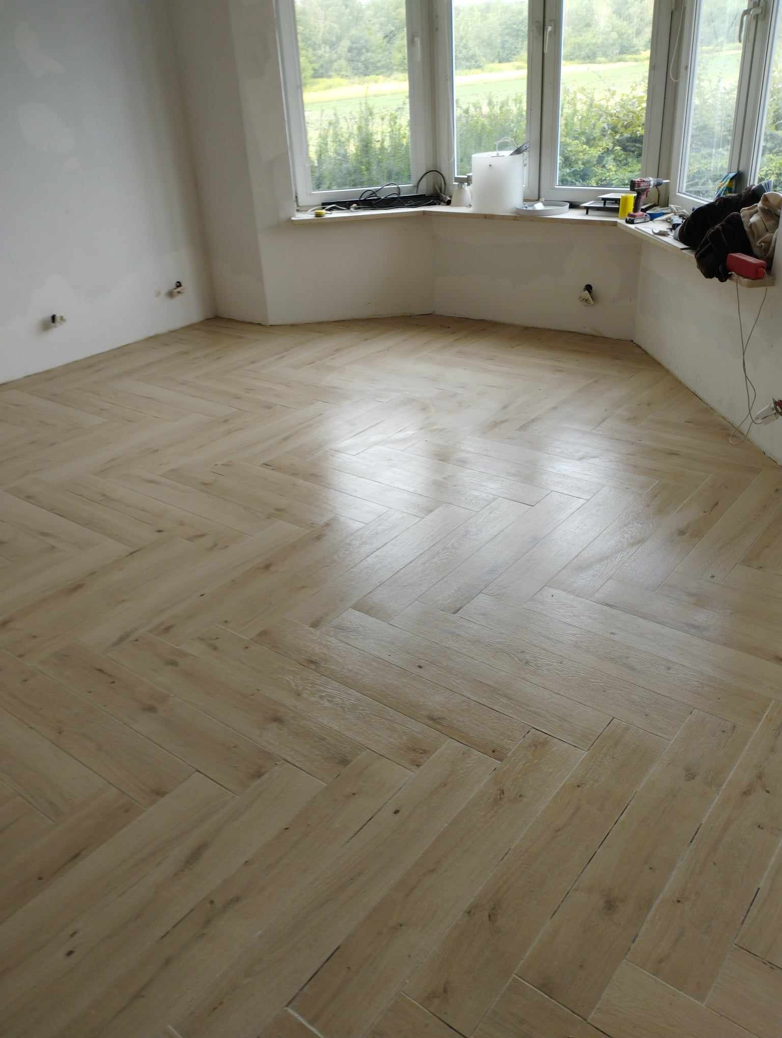 Ułożona w jodełkę, jasna, drewnopodobna podłoga z płytek w pokoju z dużym oknem i widokiem na zieleń, widoczne gniazdka elektryczne na ścianie.