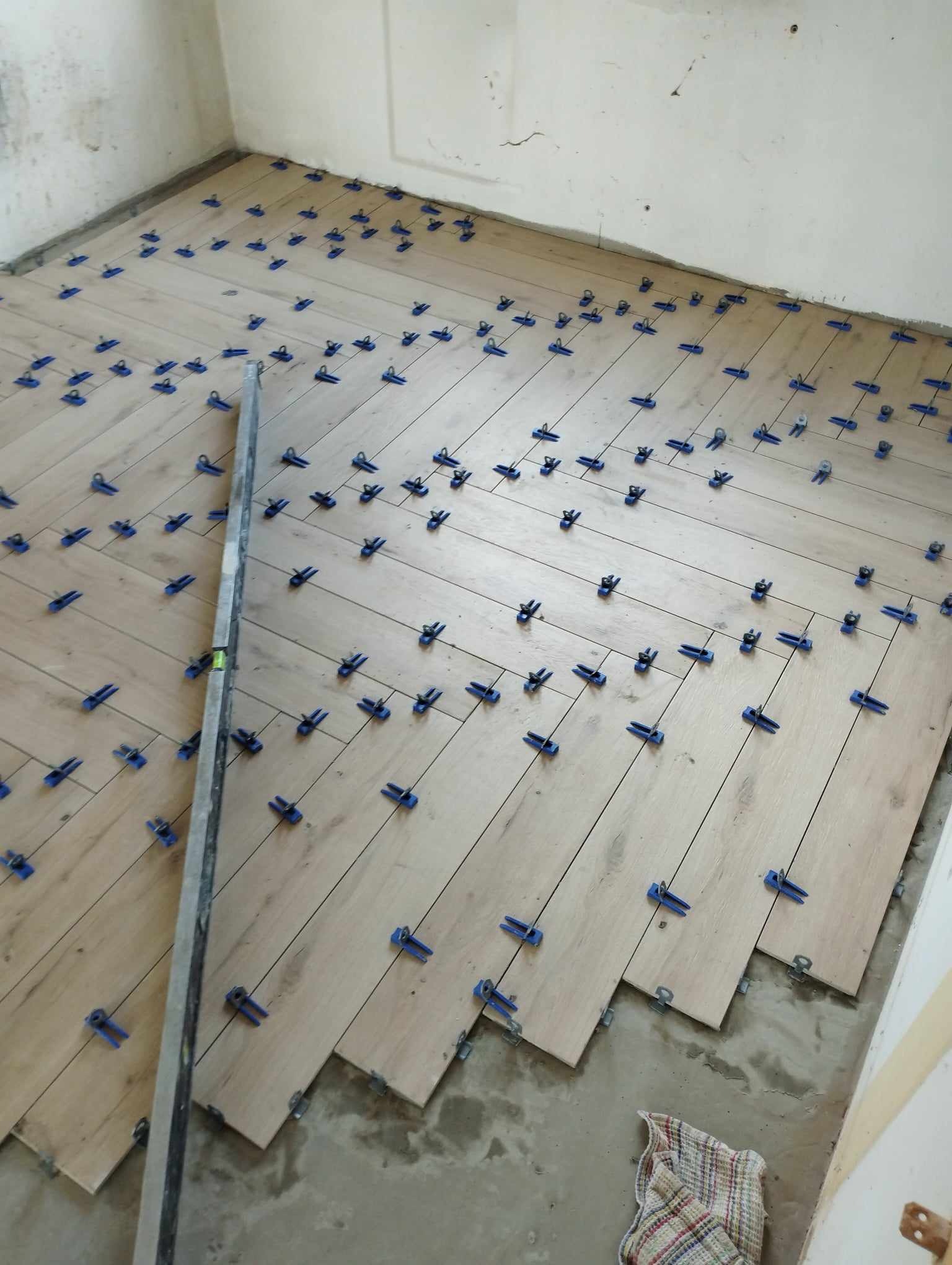 Układanie drewnopodobnych płytek podłogowych w jodełkę z użyciem niebieskich klipsów poziomujących, widoczna poziomica i fragment podłogi przed fugowaniem.