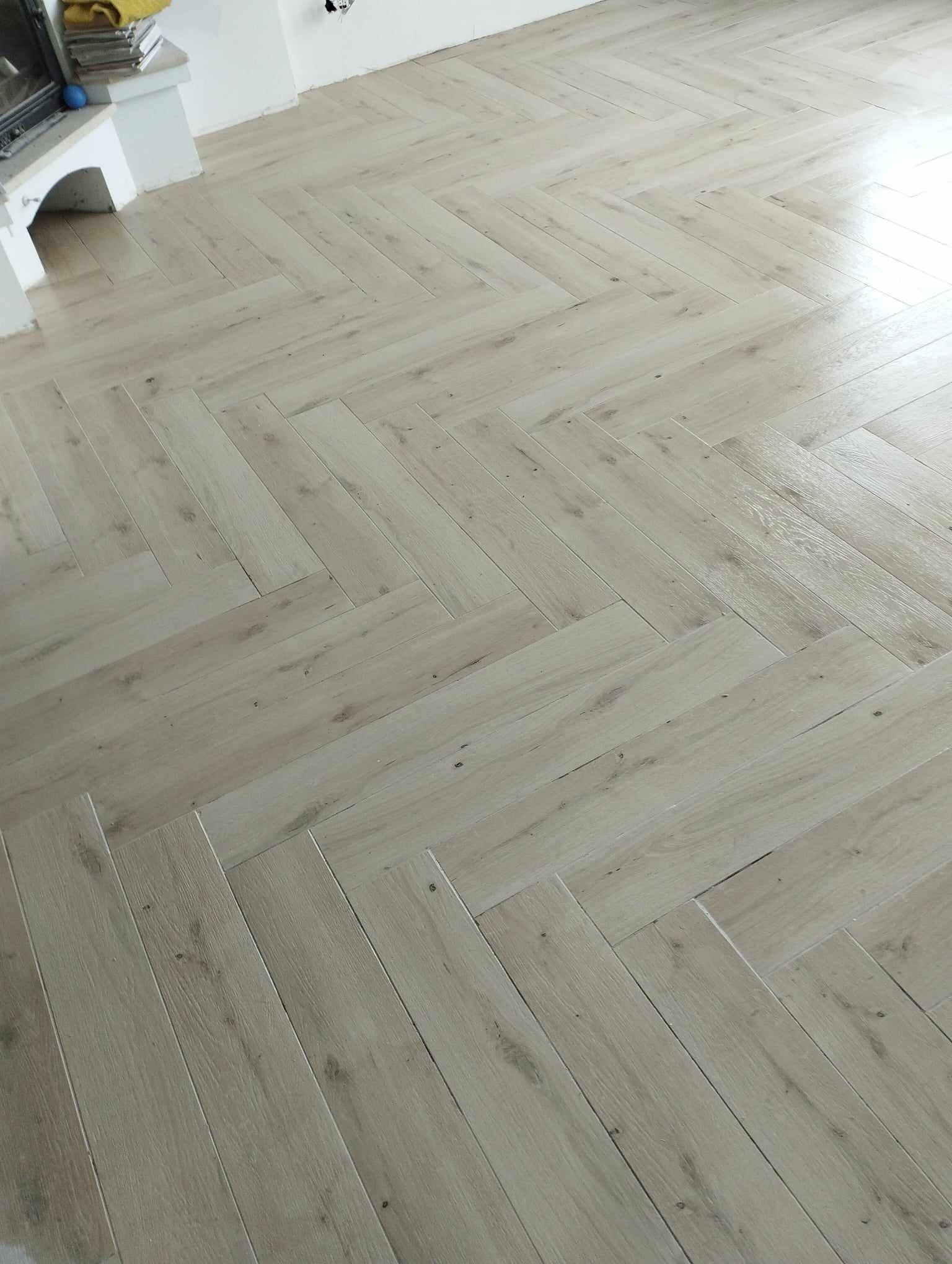 Jasna, drewnopodobna podłoga ułożona w jodełkę francuską, widoczny fragment kominka w rogu pomieszczenia.