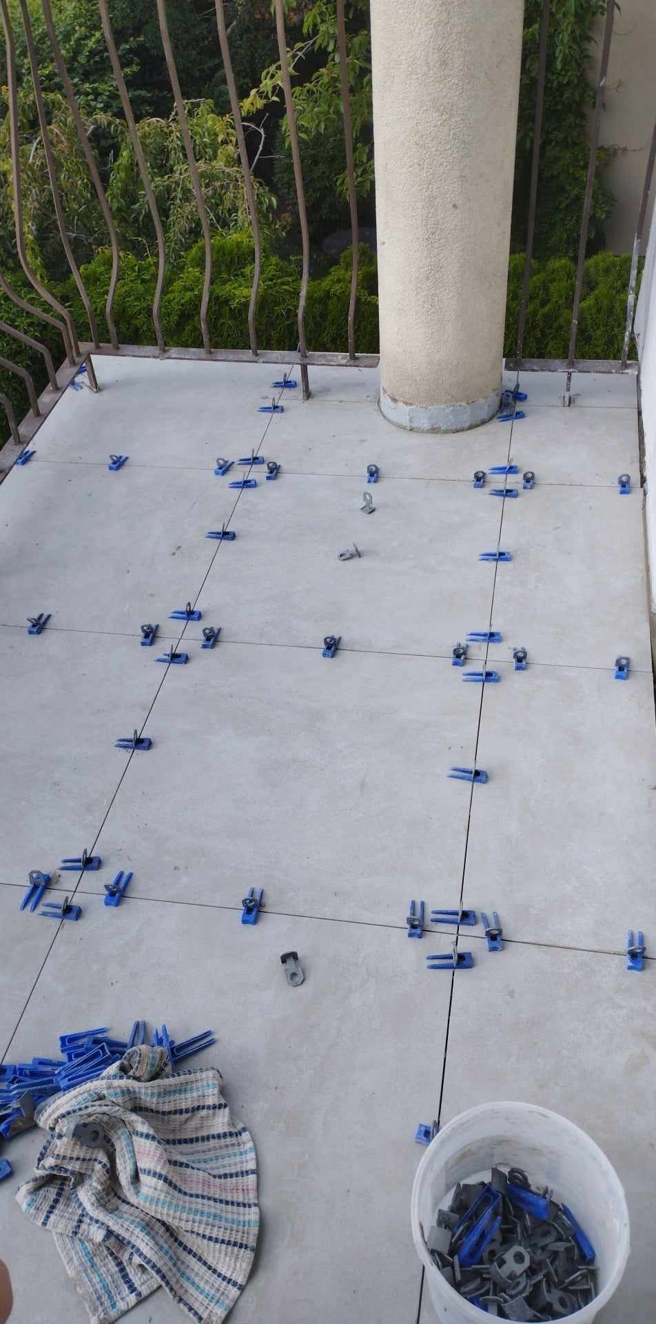 Układanie jasnych płytek na balkonie z użyciem niebieskich klipsów poziomujących i sznurków, widoczny fragment kolumny i metalowa balustrada.