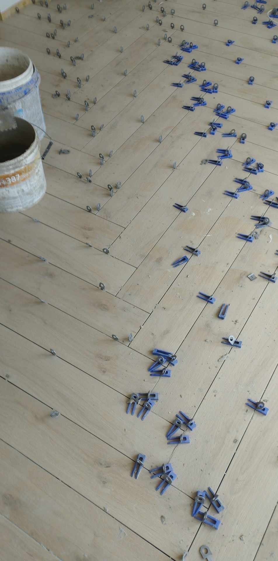 Układanie płytek podłogowych w jodełkę z użyciem niebieskich klipsów poziomujących, widoczne dwa białe wiadra po lewej stronie.