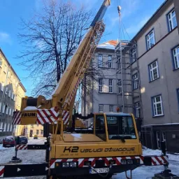 Żółty dźwig FAUN z wysięgnikiem podnosi stalową konstrukcję na tle budynku mieszkalnego w zimowy dzień.