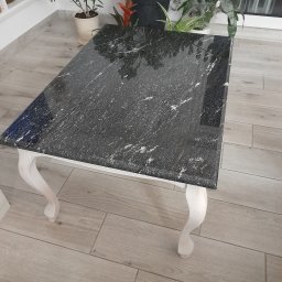 Prace Kamieniarskie Andrzej Jastrzębski - Stolik kawowy z czarnym blatem o strukturze kamienia, osadzony na białych, giętych nogach, na tle drewnopodobnej podłogi i roślin doniczkowych.