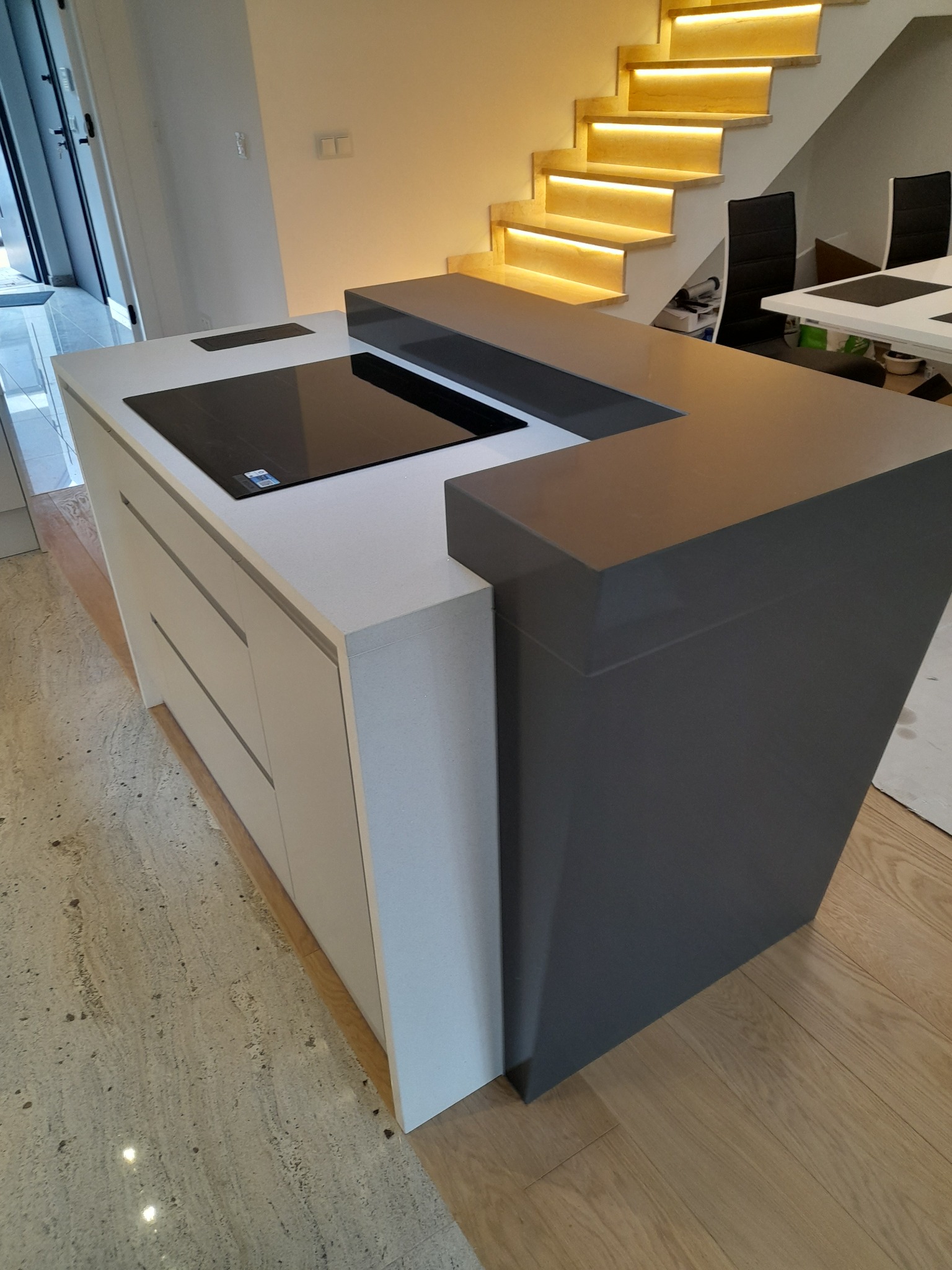 Nowoczesna wyspa kuchenna z szarym blatem i płytą indukcyjną, na tle schodów z podświetleniem LED.