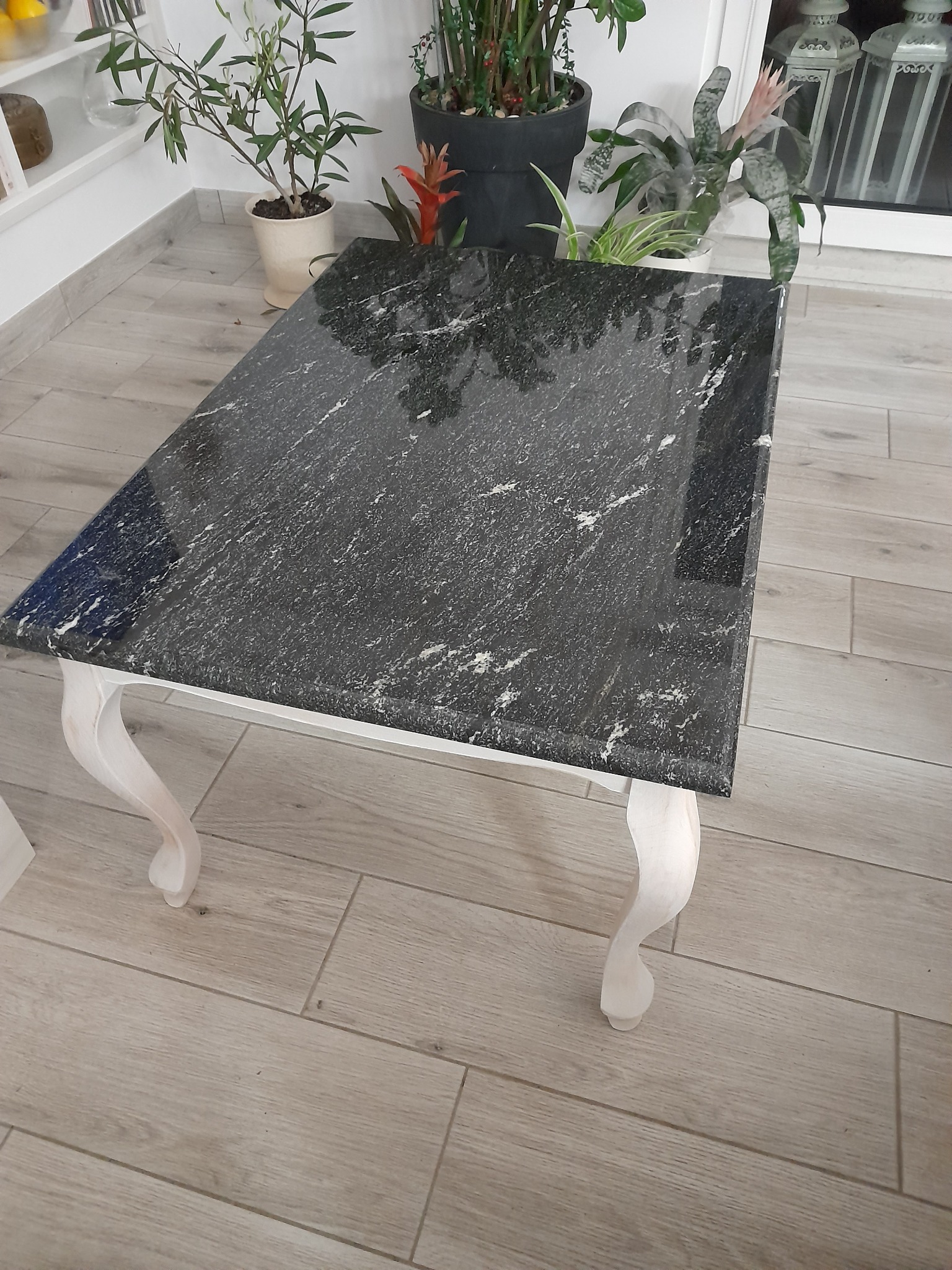 Stolik kawowy z czarnym blatem o strukturze kamienia, osadzony na białych, giętych nogach, na tle drewnopodobnej podłogi i roślin doniczkowych.