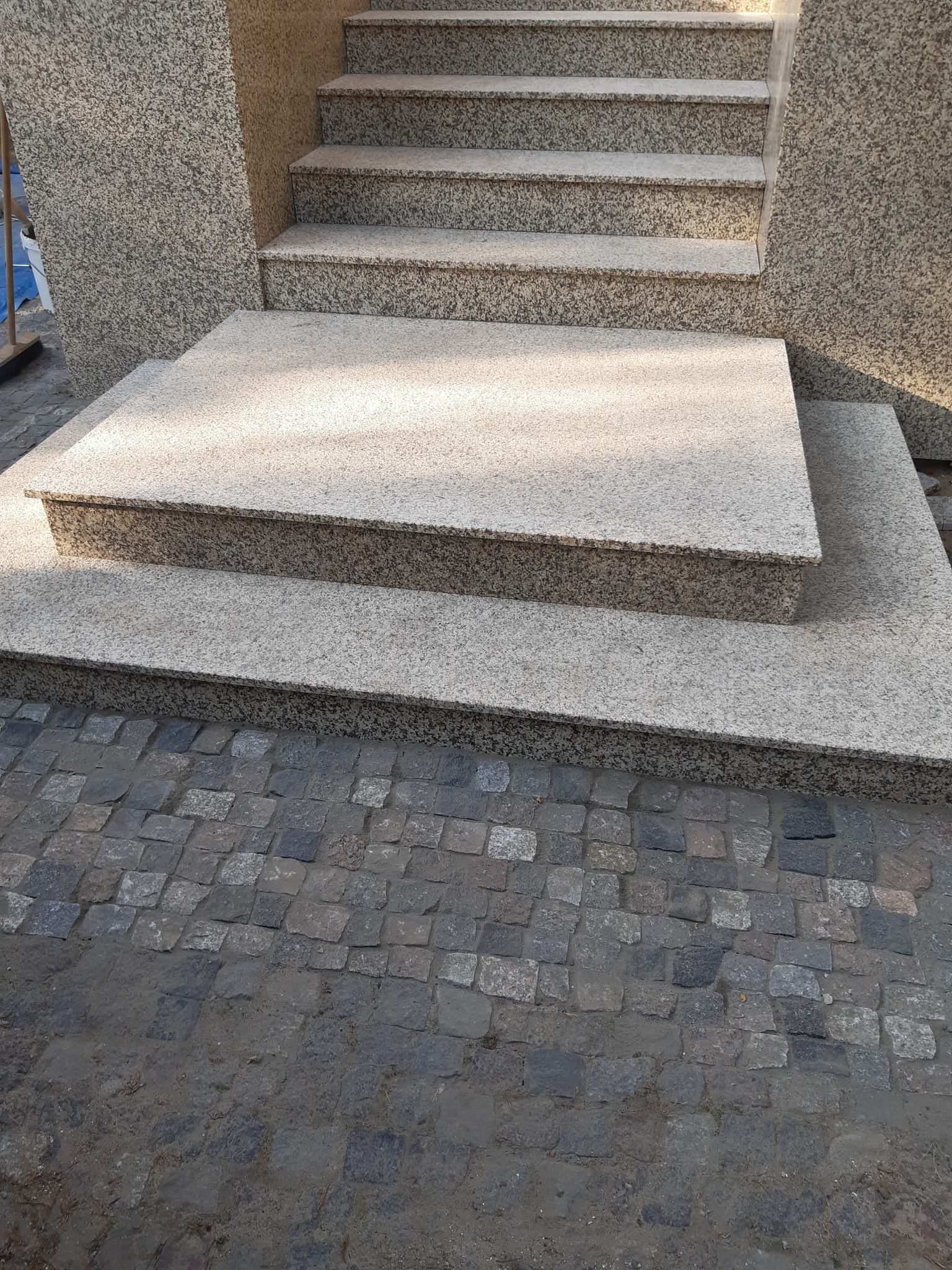 Zewnętrzne schody kamienne z granitu o jasnoszarej barwie, prowadzące do wejścia budynku, wykończone kostką brukową.