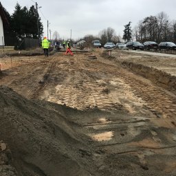 PIXEL BUD VOLODYMYR ZATSERKOVNYI SP.K. - Przygotowanie terenu pod budowę drogi: wyrównany teren z widocznymi śladami opon, pracownicy w odblaskowych ubraniach, samochody w tle, pochmurne niebo.