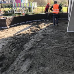 PIXEL BUD VOLODYMYR ZATSERKOVNYI SP.K. - Przygotowanie podłoża pod taras z kostki: dwóch pracowników wyrównuje piasek, w tle widoczna konstrukcja domu z betonowymi filarami oraz plac zabaw dla dzieci.