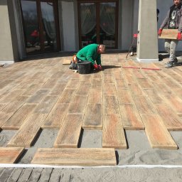 PIXEL BUD VOLODYMYR ZATSERKOVNYI SP.K. - Montaż drewnopodobnej kostki brukowej na tarasie domu, pracownik w zielonej bluzie i czerwonych rękawicach układa elementy, drugi pracownik przenosi kolejną kostkę, widoczne narzędzia...