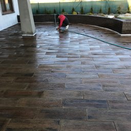 PIXEL BUD VOLODYMYR ZATSERKOVNYI SP.K. - Czyszczenie nowo ułożonej, ciemnobrązowej kostki brukowej imitującej drewno na tarasie domu jednorodzinnego, widoczny wąż ogrodowy i szczotka.