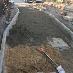 PIXEL BUD VOLODYMYR ZATSERKOVNYI SP.K. - Przygotowanie podłoża pod chodnik z obrzeżami, widoczna warstwa piasku i ziemi, w tle zaparkowane busy i budynki.